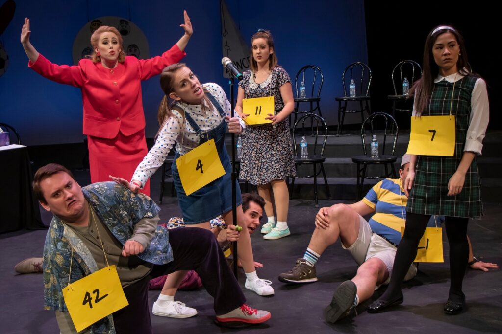 Actors in the 25th Annual Putnam County Spelling Bee posing around a mic stand. The actors playing the spellers are all in various poses as the actress portraying Rona tries to bring calm to the situation.