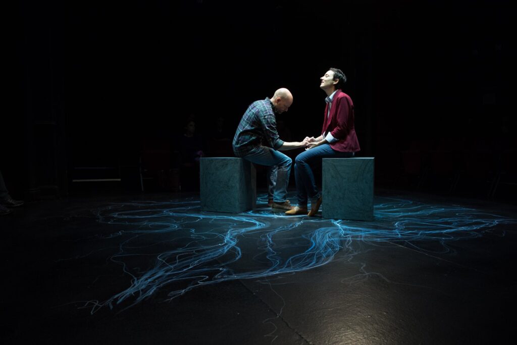 Two actors portraying the characters in the play Constellations. Both actors are seated on large black wooden cubes while holding hands.
