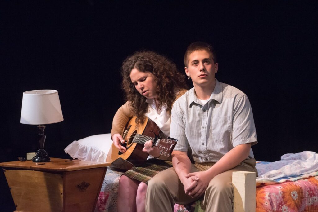 Actors portraying Rose and Birdlace in the musical Dogfight seated on a bed on stage. The actress playing rose is playing a guitar while the actor playing Eddie listens.