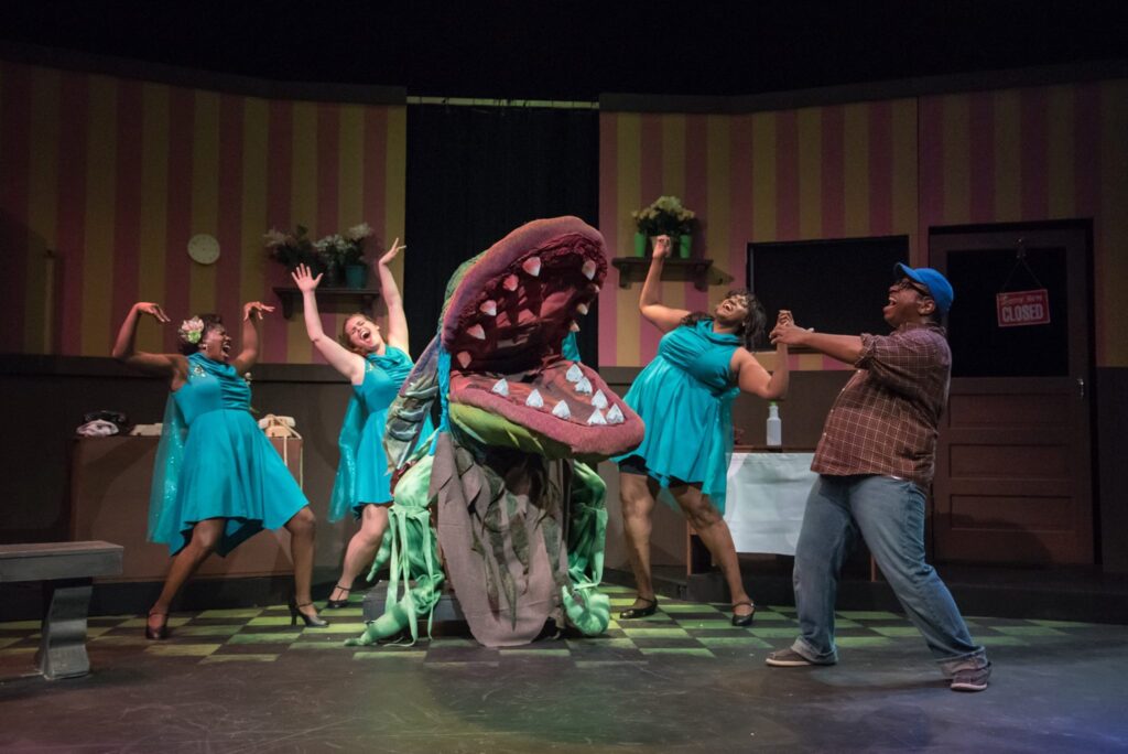 Actors playing the three urchins and Seymour in The Little Shop of Horrors singing with a large puppet of the man-eating plant Audrey II.