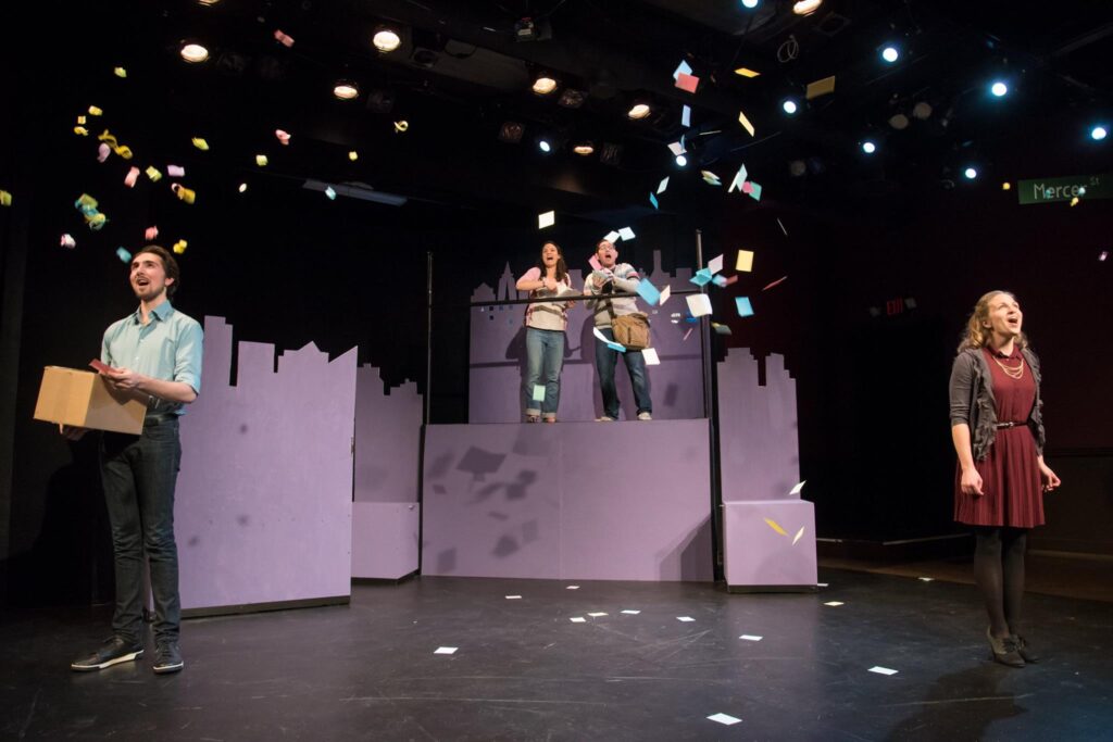 Actors in the musical Ordinary Days. Two actors in the foreground are watching as papers float down around them that were thrown by the two actors on platforms in the back.