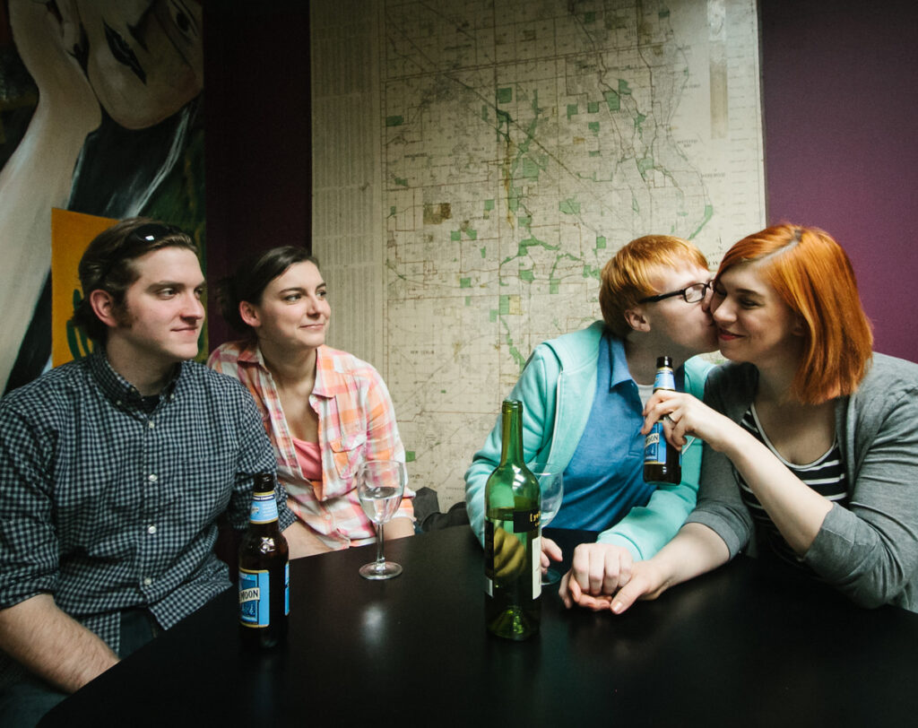 A scene from the Shape of Things with four actors portraying the main characters seated at a table. One actor is kissing another on the cheek.